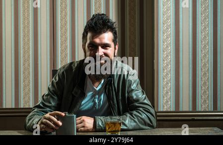 Un homme souriant et charmant qui boit seul. Hippster brutal barbu cognac cher. Problèmes graves de dépendance à l'alcool. Alcoolisme Banque D'Images