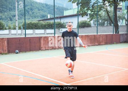 Jeune homme avec un chignon homme jongle un ballon de football sur un terrain orange vibrant pendant une journée ensoleillée. Portant des vêtements de sport décontractés, il se concentre sur le contrôle du bal Banque D'Images