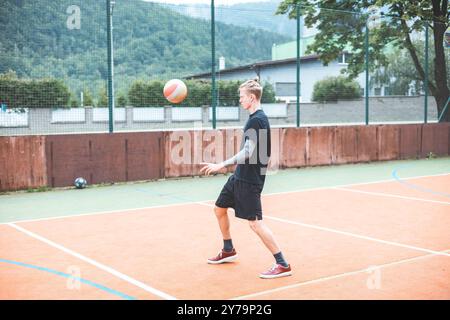 Jeune homme avec un chignon homme jongle un ballon de football sur un terrain orange vibrant pendant une journée ensoleillée. Portant des vêtements de sport décontractés, il se concentre sur le contrôle du bal Banque D'Images