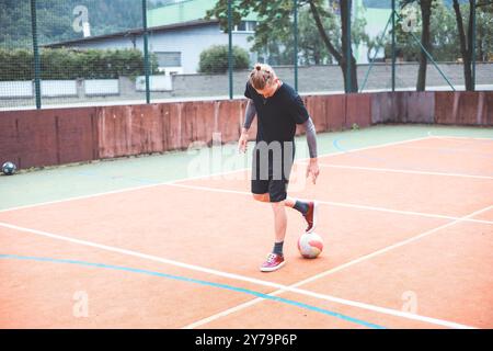 Jeune homme avec un chignon homme jongle un ballon de football sur un terrain orange vibrant pendant une journée ensoleillée. Portant des vêtements de sport décontractés, il se concentre sur le contrôle du bal Banque D'Images