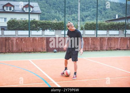 Jeune homme avec un chignon homme jongle un ballon de football sur un terrain orange vibrant pendant une journée ensoleillée. Portant des vêtements de sport décontractés, il se concentre sur le contrôle du bal Banque D'Images