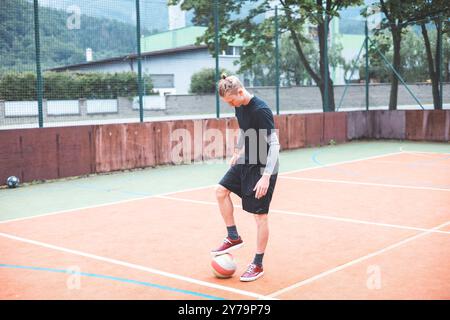 Jeune homme avec un chignon homme jongle un ballon de football sur un terrain orange vibrant pendant une journée ensoleillée. Portant des vêtements de sport décontractés, il se concentre sur le contrôle du bal Banque D'Images