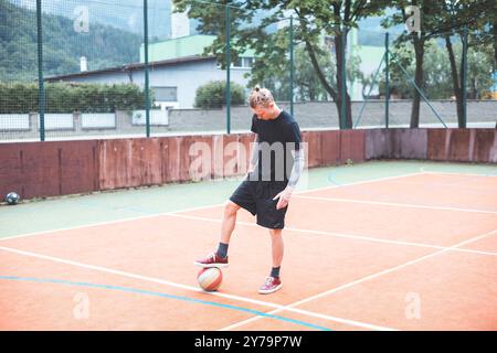 Jeune homme avec un chignon homme jongle un ballon de football sur un terrain orange vibrant pendant une journée ensoleillée. Portant des vêtements de sport décontractés, il se concentre sur le contrôle du bal Banque D'Images