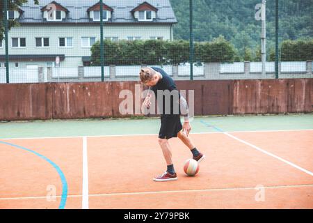 Jeune homme avec un chignon homme jongle un ballon de football sur un terrain orange vibrant pendant une journée ensoleillée. Portant des vêtements de sport décontractés, il se concentre sur le contrôle du bal Banque D'Images