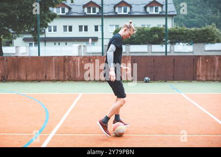 Jeune homme avec un chignon homme jongle un ballon de football sur un terrain orange vibrant pendant une journée ensoleillée. Portant des vêtements de sport décontractés, il se concentre sur le contrôle du bal Banque D'Images