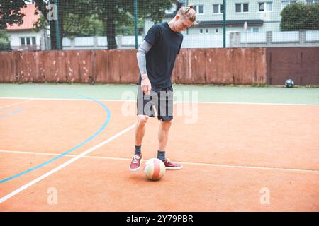 Jeune homme avec un chignon homme jongle un ballon de football sur un terrain orange vibrant pendant une journée ensoleillée. Portant des vêtements de sport décontractés, il se concentre sur le contrôle du bal Banque D'Images