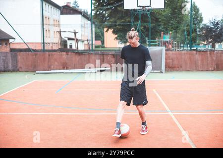 Jeune homme avec un chignon homme jongle un ballon de football sur un terrain orange vibrant pendant une journée ensoleillée. Portant des vêtements de sport décontractés, il se concentre sur le contrôle du bal Banque D'Images