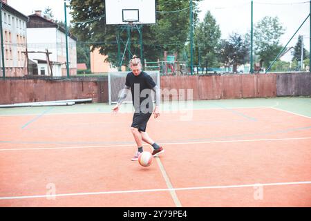 Jeune homme avec un chignon homme jongle un ballon de football sur un terrain orange vibrant pendant une journée ensoleillée. Portant des vêtements de sport décontractés, il se concentre sur le contrôle du bal Banque D'Images