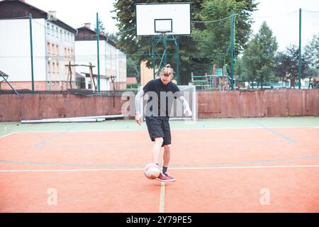 Jeune homme avec un chignon homme jongle un ballon de football sur un terrain orange vibrant pendant une journée ensoleillée. Portant des vêtements de sport décontractés, il se concentre sur le contrôle du bal Banque D'Images