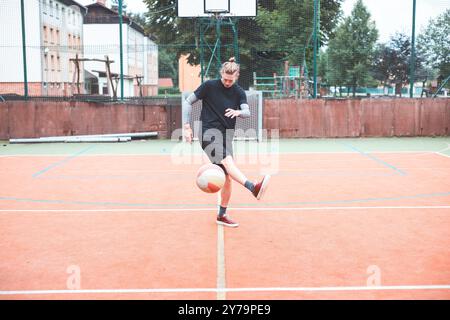 Jeune homme avec un chignon homme jongle un ballon de football sur un terrain orange vibrant pendant une journée ensoleillée. Portant des vêtements de sport décontractés, il se concentre sur le contrôle du bal Banque D'Images