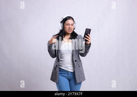 belle fille de bureau asie ennuyé de gadget portant casque et tenant téléphone portable tout en pointant le costume portant isolé sur fond blanc. la vie Banque D'Images