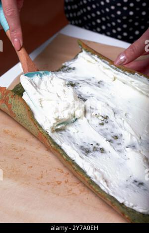 Une femme met de la crème sur le gâteau pour préparer un biscuit roulé avec des épinards et de la menthe. Farci aux fraises. Ingrédients pour la cuisson. Banque D'Images