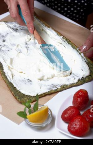 Une femme met de la crème sur le gâteau pour préparer un biscuit roulé avec des épinards et de la menthe. Farci aux fraises. Ingrédients pour la cuisson. Banque D'Images