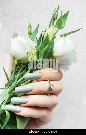 Une main avec de longs ongles élégants tient un bouquet de fleurs blanches fraîches. Les verts et les blancs doux contrastent magnifiquement avec un dos texturé léger Banque D'Images