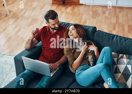 Vue de dessus de jeune couple heureux dans des vêtements décontractés se liant avec chat domestique et souriant tout en se reposant sur le canapé à l'intérieur Banque D'Images