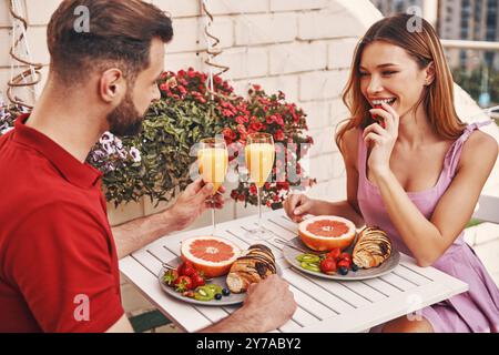 Mignon jeune couple dans des vêtements décontractés prenant le petit déjeuner et souriant tout en étant assis sur le patio sur le toit à l'extérieur Banque D'Images