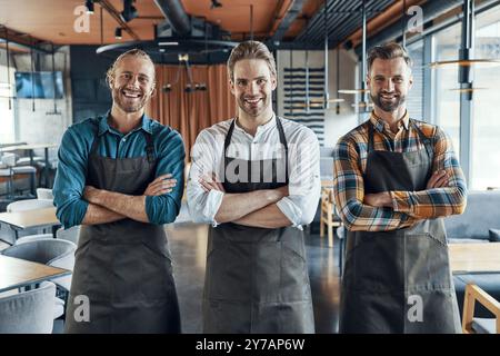 Trois beaux serveurs masculins dans des tabliers gardant les bras croisés et regardant la caméra avec le sourire tout en se tenant debout dans le restaurant Banque D'Images