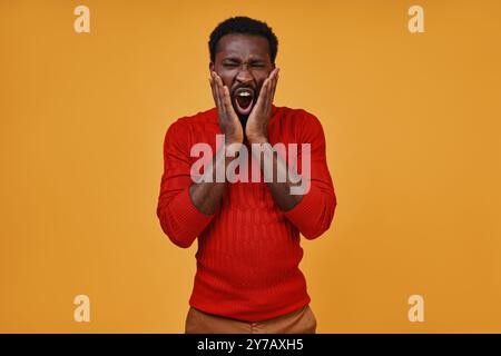 Frustré jeune homme africain dans des vêtements décontractés tenant le visage dans les mains tout en se tenant debout sur fond jaune Banque D'Images