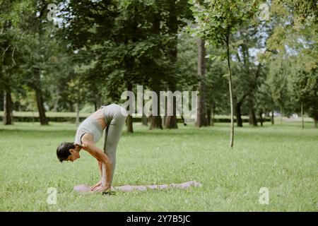 Femme senior vêtue de vêtements de sport vert pastel pratiquant le yoga dans le parc verdoyant local Banque D'Images
