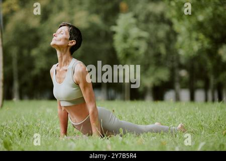 Femme senior immergée dans les pensées tout en se tenant debout dans la position Urdhva Mukha Svanasana sur la pelouse fraîche coupée Banque D'Images
