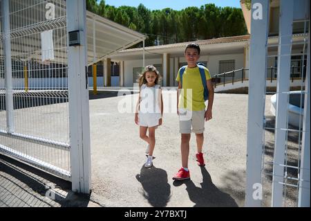 Deux jeunes élèves avec des sacs à dos traversent les portes de l'école, capturant l'essence d'une journée d'école ensoleillée. Banque D'Images