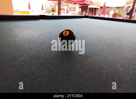 Gros plan d'une boule de billard noire numéro 6 placée sur une table de billard dans un cadre extérieur. Le fond montre des meubles de patio flous et des parasols, Banque D'Images