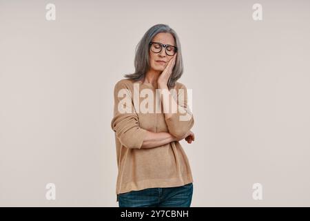 Femme mature frustrée souffrant de maux de dents et touchant sa joue tout en se tenant debout sur fond gris Banque D'Images