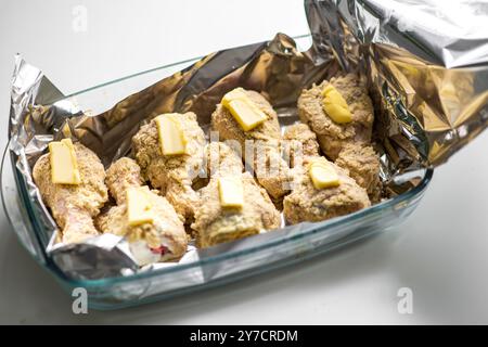 Pilons de poulet crus panés avec beurre dans un plat de cuisson en verre recouvert de papier d'aluminium. Banque D'Images