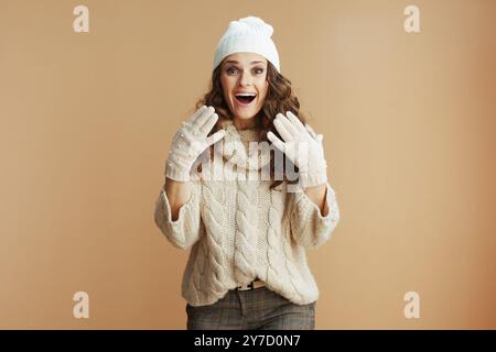 Bonjour l'hiver. Portrait de la femme élégante surprise de 40 ans en pull beige, mitaines et chapeau isolé sur fond beige. Banque D'Images
