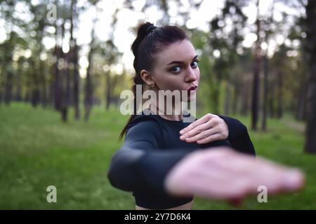 Jeune femme faisant la formation d'arts martiaux dans le haut noir sportif tenant les mains dans la pose de combat sur fond de forêt Banque D'Images