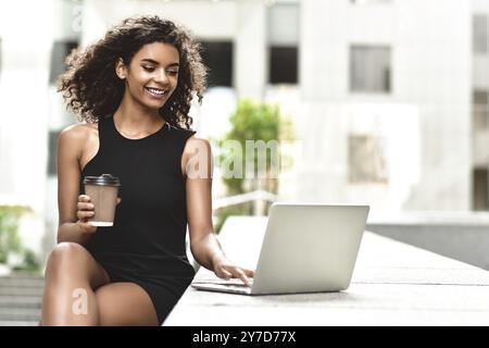 Jeune fille mixte attrayante avec de beaux cheveux afro qui sourit en regardant l'écran de son ordinateur portable pendant l'appel vidéo. Jeune femme utilisant un ordinateur portable Banque D'Images