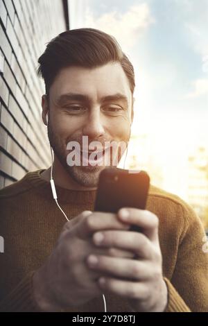 Smiling man listening to music on headphones et appuyé contre un mur de briques. Portrait of smiling man avec des écouteurs et un comité permanent par brique w Banque D'Images