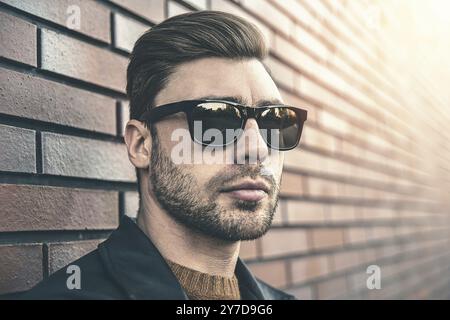 Portrait d'élégant beau jeune homme avec des poils debout à l'extérieur et appuyé sur le mur de briques. Homme portant une veste, un pull et des lunettes de soleil Banque D'Images