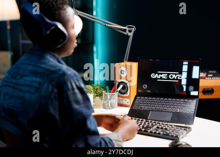 Femme afro-américaine avec des écouteurs est assis devant son ordinateur personnel qui affiche le jeu terminé. Joueuse portant un casque, perdant un jeu est regarder son écran numérique d'ordinateur portable. Banque D'Images