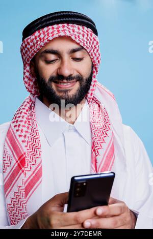 Homme arabe souriant portant des vêtements traditionnels islamiques tapant un message sur smartphone. Personne musulmane vêtue de thobe et de ghutra discutant dans les médias sociaux en utilisant un téléphone portable avec une expression joyeuse Banque D'Images