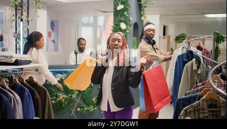 Couple afro-américain s'amusant dans la boutique de mode, faisant des mouvements de danse. Les clients joyeux s'amusent à la musique de magasin de vêtements de centre commercial agréable, dansant avec des sacs à provisions Banque D'Images