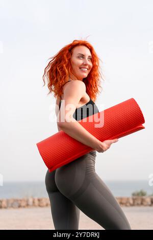 Joyeux jogger femme tenant le tapis d'exercice enroulé contre le ciel clair. La vue latérale de la jeune femme en tenue de sport est détournée pendant la journée. Banque D'Images