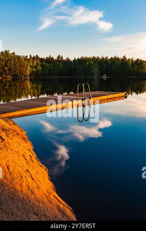 Un lac serein en Suède dispose d'une jetée en bois s'étendant dans les eaux calmes. Le coucher de soleil jette de beaux reflets, améliorant l'atmosphère paisible Banque D'Images