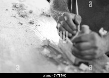 Gros plan des mains du charpentier à l'aide d'un rabot à main pour lisser une planche de bois dans son atelier. - noir n blanc - Banque D'Images