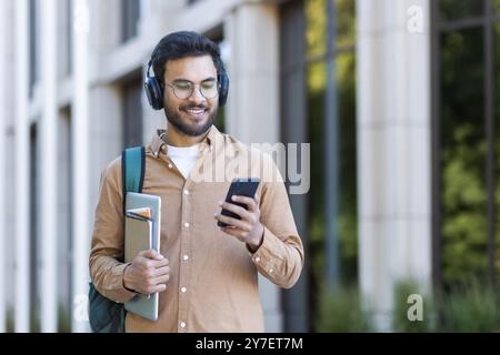 Jeune homme confiant debout dehors portant des écouteurs et tenant le téléphone. Sourire, consulter les messages et écouter de la musique. Style décontracté avec chemise marron, tenant le cahier et la tablette. Banque D'Images