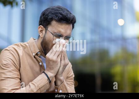 Jeune homme portant des lunettes et des vêtements décontractés éternue dans le tissu à l'extérieur de l'immeuble de bureaux moderne. L'homme montre des signes d'allergies saisonnières, mettant en évidence les défis de santé dans les environnements urbains. Banque D'Images