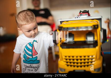 Un enfant en bas âge examine un grand camion jaune avec curiosité, entouré de jouets colorés dans un salon chaleureux et accueillant tandis qu'un parent se détend en t Banque D'Images