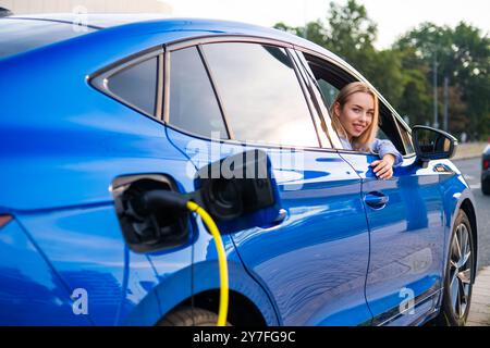 Femme blonde regarde par la fenêtre de la voiture pendant que son véhicule électrique se recharge à un point de recharge pratique à l'extérieur Banque D'Images