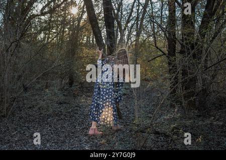 fille dans une robe fleurie explore une forêt ensoleillée, tenant sur un arbre Banque D'Images