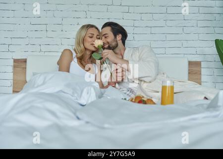 Mignon couple aimant sentant rose tout en prenant le petit déjeuner au lit Banque D'Images