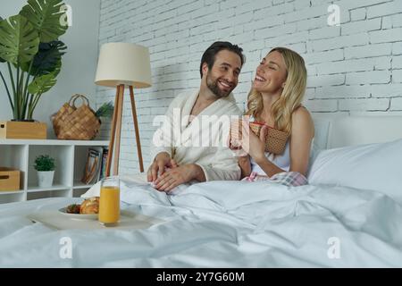 Femme heureuse recevant une boîte cadeau de ses petits amis alors que les deux sont assis dans le lit Banque D'Images