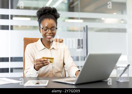 Femme d'affaires confiante souriant tout en faisant l'achat en ligne à l'aide de la carte de crédit et ordinateur portable dans un bureau. L'image représente la commodité, la technologie et l'environnement professionnel Banque D'Images