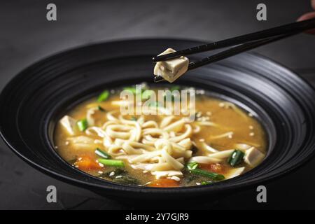 Les baguettes tiennent un morceau de fromage tofu sur une assiette de soupe miso avec des nouilles udon, du poulet, des oignons verts et des graines de sésame dans un bol noir avec du chopst Banque D'Images