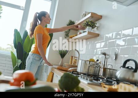 Jeune femme confiante mettant des assiettes sur l'étagère tout en se tenant debout à la cuisine domestique Banque D'Images