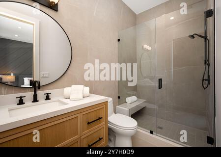 Salle de bains moderne avec cabine de douche en verre, meuble-lavabo en bois et grand miroir ovale aux tons neutres et aux luminaires contemporains. Banque D'Images